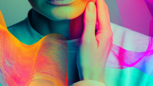 Eine künstlerische Nahaufnahme zur verdeutlichung des Themas "Stimmtransition bei trans* Personen"mit lebendigen Neonfarben. Eine Hand berührt nachdenklich das Kinn. Der untere Teil des Gesichts ist sichtbar, während der Rest außerhalb des Bildrahmens liegt. Die gesamte Szene ist in leuchtenden Farbtönen von Türkis, Pink, Orange und Grün beleuchtet. Wellenförmige, farbige Lichtmuster überlagern das Bild und erzeugen einen surrealen, digitalen Kunsteffekt. Die Ästhetik erinnert an moderne digitale Kunst oder Vaporwave-Stil mit starken Farbkontrasten und einer traumhaften Atmosphäre.
